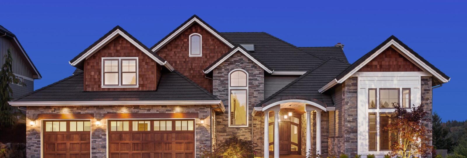 Two-story house with stone and wood exterior, illuminated by exterior lights. The home has a double garage and several windows. The sky is clear and blue.